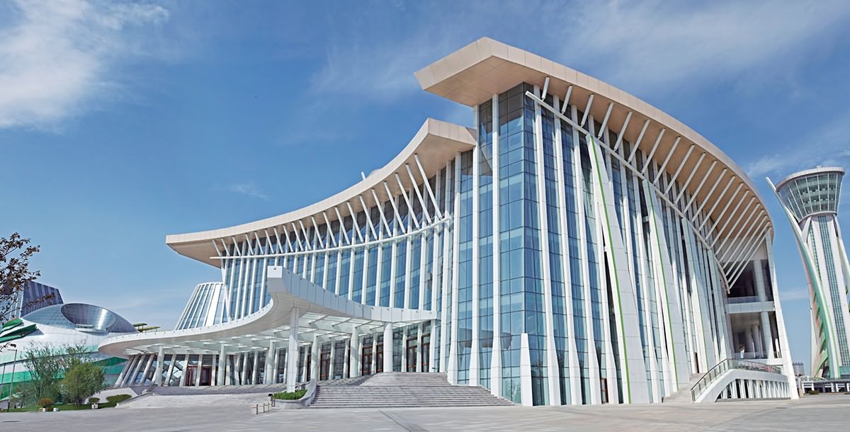 Modern curved glass and metal building exterior with sweeping canopy roof under a blue sky, white and beige tones