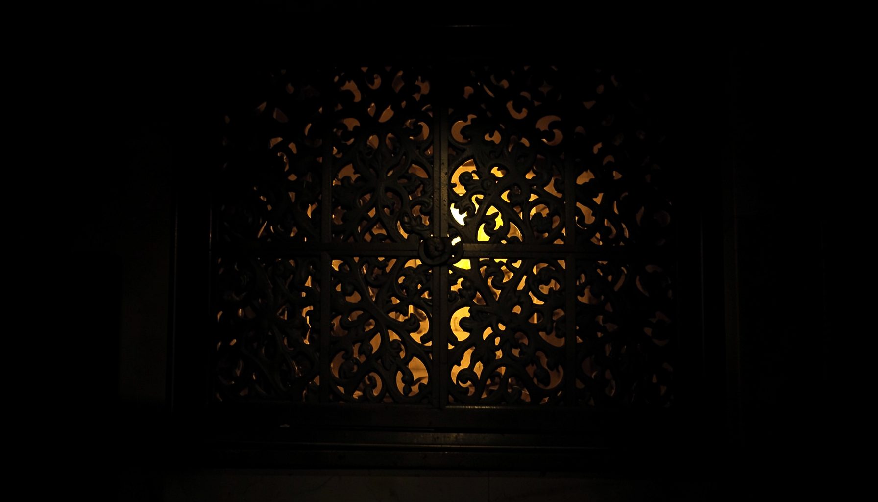 Dark architectural interior with an ornate metal carved panel featuring intricate cut-out patterns, illuminated by warm light
