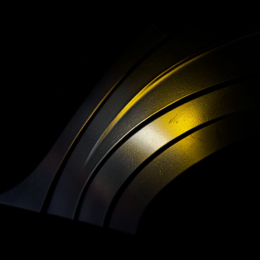 Dark curved aluminum panel with sculpted double-curved carving under dramatic side lighting revealing deep relief
