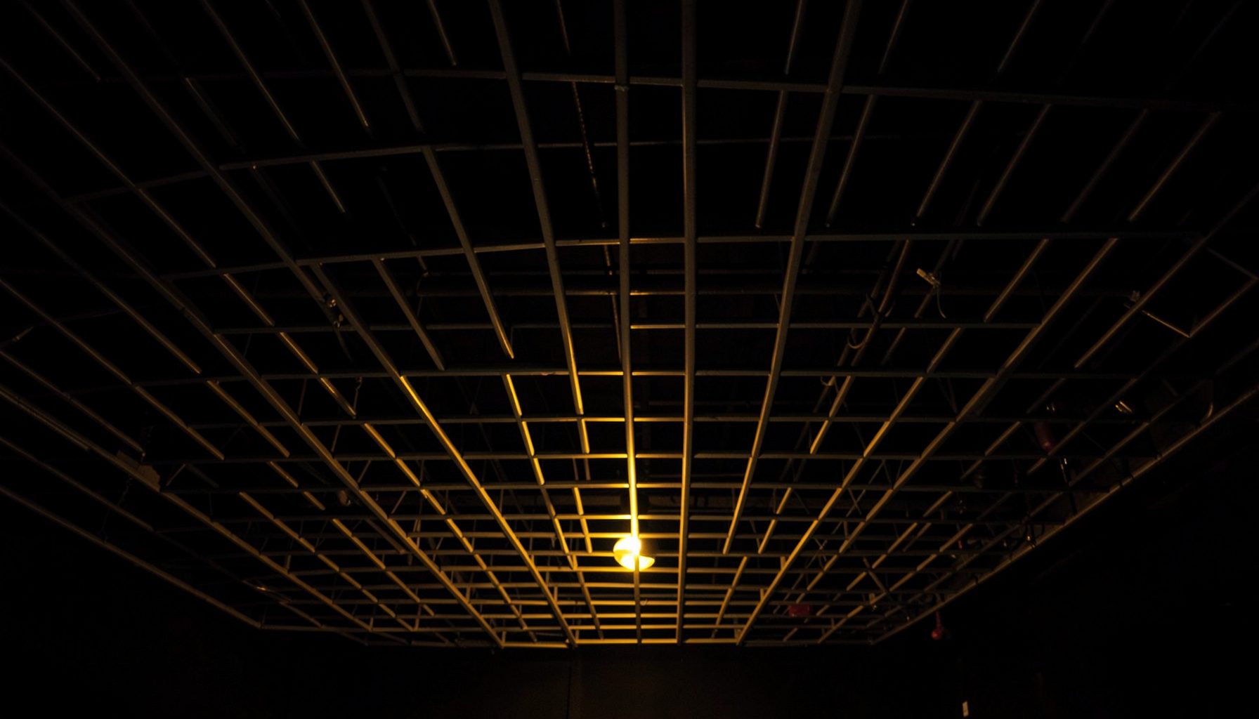Dark interior ceiling with rows of suspended aluminum square tubes creating a linear open-grid pattern, warm ambient lighting